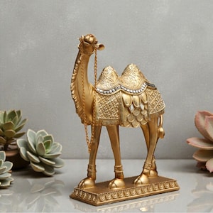 Op de afbeelding: Een goudkleurig kameelbeeldje met ingewikkelde details. De decoratieve kameel heeft een zadel, een teugel en een ketting. Het beeldje staat op een rechthoekige basis met een sierlijke rand. Het is een decoratief item.