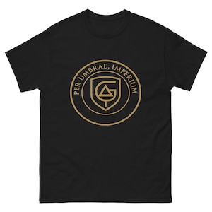 Puede incluir: Camiseta negra con un emblema dorado. El emblema presenta el texto "PER UMBRAE, IMPERIUM" alrededor de un escudo con un diseño estilizado de "G" y triángulo. La camiseta es de cuello redondo.