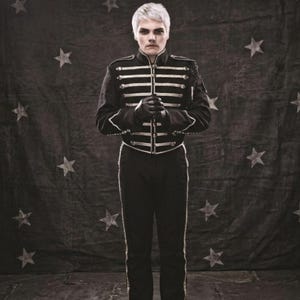 May include: A person with short platinum blonde hair wearing a black military-style jacket featuring gold-toned horizontal braided trim and matching black trousers with a thin gold stripe down the side, standing against a dark star-patterned backdrop.