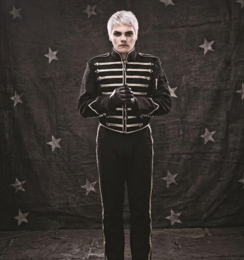 May include: A person with short platinum blonde hair wearing a black military-style jacket featuring gold-toned horizontal braided trim and matching black trousers with a thin gold stripe down the side, standing against a dark star-patterned backdrop.