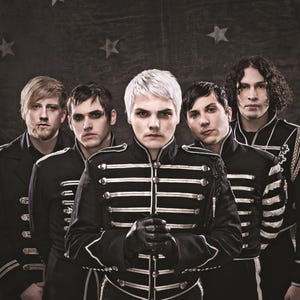 May include: Five individuals stand in a row wearing black military-style jackets with silver piping and buttons. The central figure has platinum blonde hair and black gloves. They are positioned against a dark backdrop featuring a star pattern.