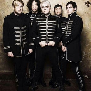 Puede incluir: Un grupo de cinco personas posan con chaquetas negras de estilo militar con adornos y detalles plateados. Las chaquetas presentan múltiples filas de rayas y botones plateados. El fondo es una pared texturizada en tonos sepia. El grupo probablemente sea una banda o un conjunto musical.