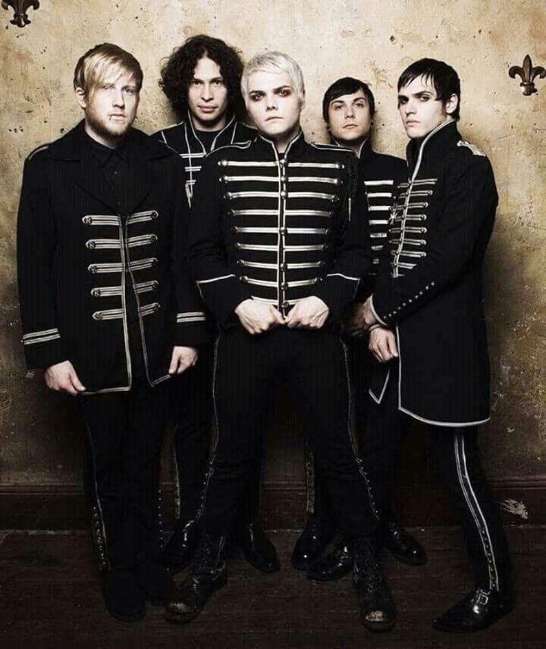 May include: Five individuals standing in a row wearing black military-style jackets with silver horizontal braided trim and matching black trousers. The jackets feature high collars and structured silhouettes, creating a uniform appearance across the group.