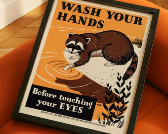 Puede incluir: Un póster vintage enmarcado con un mapache lavándose las manos, sobre un fondo naranja y amarillo. El póster dice: "Lávate las manos antes de tocarte los ojos". El marco es negro y el póster está sobre un sofá naranja.