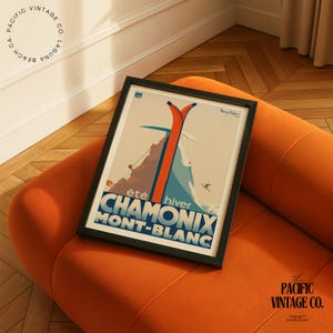 May include: A framed vintage travel poster for Chamonix Mont-Blanc, featuring orange skis, a mountain scene, and the text "&eacute;t&eacute; hiver CHAMONIX MONT-BLANC." The poster is in a black frame and rests on an orange sofa. The Pacific Vintage Co. logo is visible.