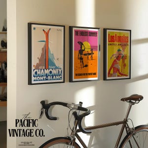 May include: Three framed vintage travel posters hang on a white wall. The posters feature retro designs with text that reads "CHAMONIX MONT-BLANC", "THE ENDLESS SUMMER", and "Los Angeles UNITED AIR LINES". A brown bicycle is in the foreground.