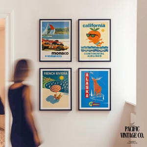 May include: Four framed vintage travel posters. The posters feature retro designs for Monaco, California, French Riviera, and Alaska. Each poster has a black frame and is mounted on a white wall. The California poster features an orange with sunglasses.