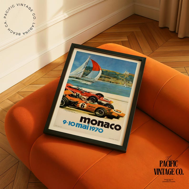 May include: A framed vintage poster featuring race cars speeding along a coastal road with a sailboat in the background. The poster has the text "Monaco 9-10 mai 1970" and is set against an orange background.