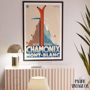 May include: A framed vintage travel poster for Chamonix Mont-Blanc, featuring orange skis, a skier, and the text "&eacute;t&eacute; hiver". The poster is 24x36 inches and is displayed above a white cabinet with a gold lamp.
