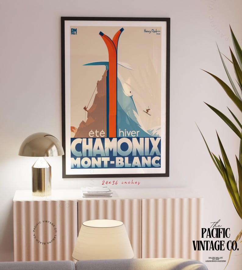 May include: A framed vintage travel poster for Chamonix Mont-Blanc, featuring orange skis, a skier, and the text "&eacute;t&eacute; hiver". The poster is 24x36 inches and is displayed above a white cabinet with a gold lamp.