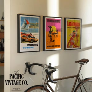 May include: Three framed vintage travel posters hang on a white wall. The posters feature retro designs with text that reads "Monaco", "The Endless Summer", and "Los Angeles". A brown bicycle is in the foreground.