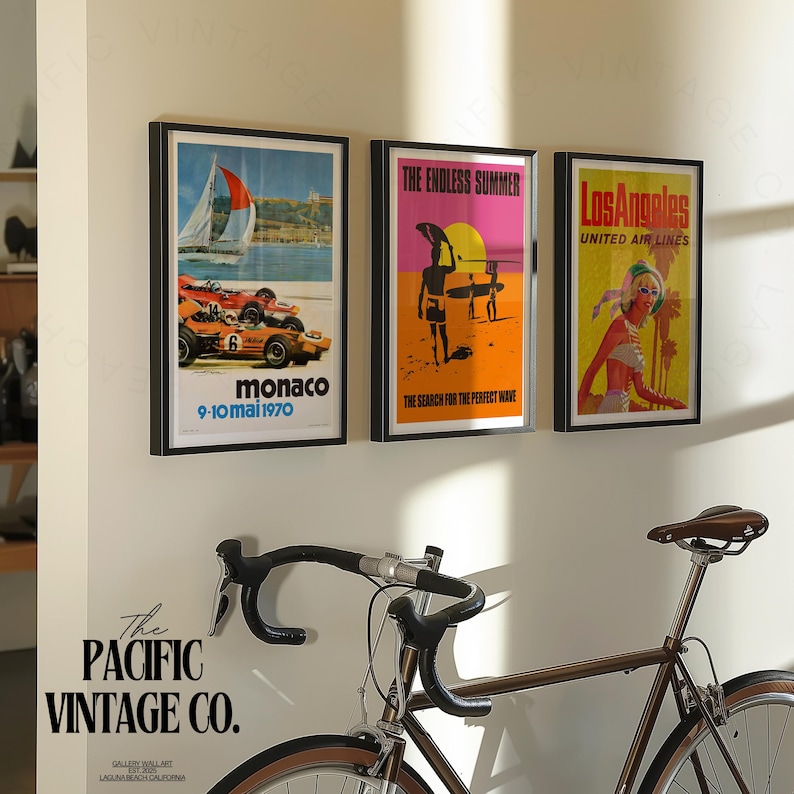 May include: Three framed vintage travel posters hang on a white wall. The posters feature retro designs with text that reads "Monaco", "The Endless Summer", and "Los Angeles". A brown bicycle is in the foreground.