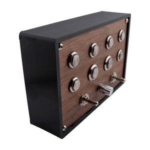 Puede incluir: Una caja rectangular negra con un panel frontal de imitación madera. El panel presenta diez botones plateados y tres interruptores de palanca. Probablemente sea un dispositivo electrónico o un panel de control.