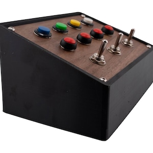 Könnte beinhalten: Eine schwarze Box mit einer Holzplatte, die farbige Knöpfe und silberne Kippschalter aufweist. Die Knöpfe sind blau, grün, gelb, weiß und rot. Die Schalter sind in einer Reihe angeordnet. Die Box ist abgewinkelt.