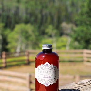 May include: A round amber-colored plastic bottle with a silver screw-top cap. The bottle features a white decorative label with the text Ranch Organics and lavender printed in a dark font, centered on the front of the container.