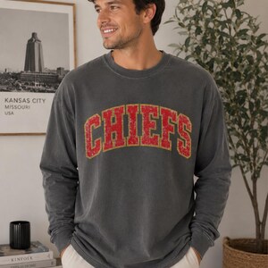 May include: A charcoal grey long-sleeve crewneck sweatshirt featuring the word CHIEFS in a distressed red font with a yellow outline arched across the chest. The garment has a relaxed fit with ribbed cuffs and a ribbed neckline.