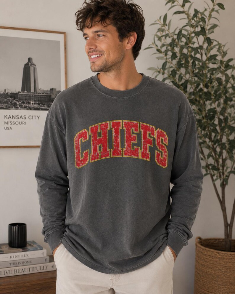 May include: A charcoal grey long-sleeve crewneck sweatshirt featuring the word CHIEFS in a distressed red font with a yellow outline arched across the chest. The garment has a relaxed fit with ribbed cuffs and a ribbed neckline.
