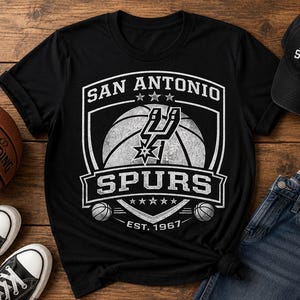May include: Black t-shirt with a white San Antonio Spurs logo featuring a basketball and the text "SPURS". A black baseball cap with the Spurs logo and a basketball are also visible. The shirt and cap are on a wooden surface.