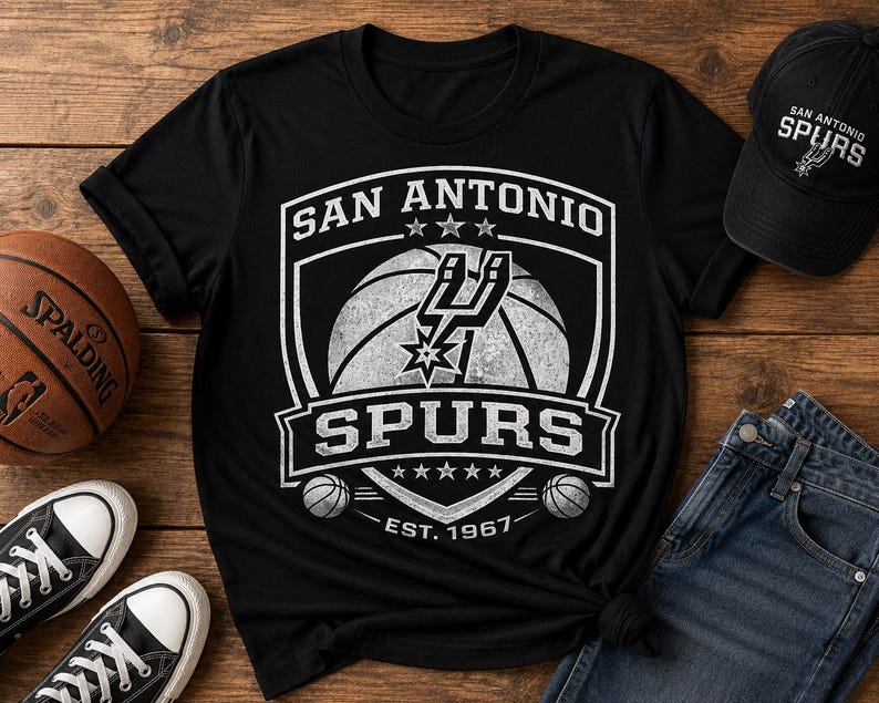 May include: Black t-shirt with a white San Antonio Spurs logo featuring a basketball and the text "SPURS". A black baseball cap with the Spurs logo and a basketball are also visible. The shirt and cap are on a wooden surface.