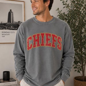 May include: A charcoal grey long-sleeve crewneck sweatshirt featuring the word CHIEFS printed in a distressed red font with a gold outline across the chest. The garment has a relaxed fit with ribbed cuffs and a ribbed neckline.