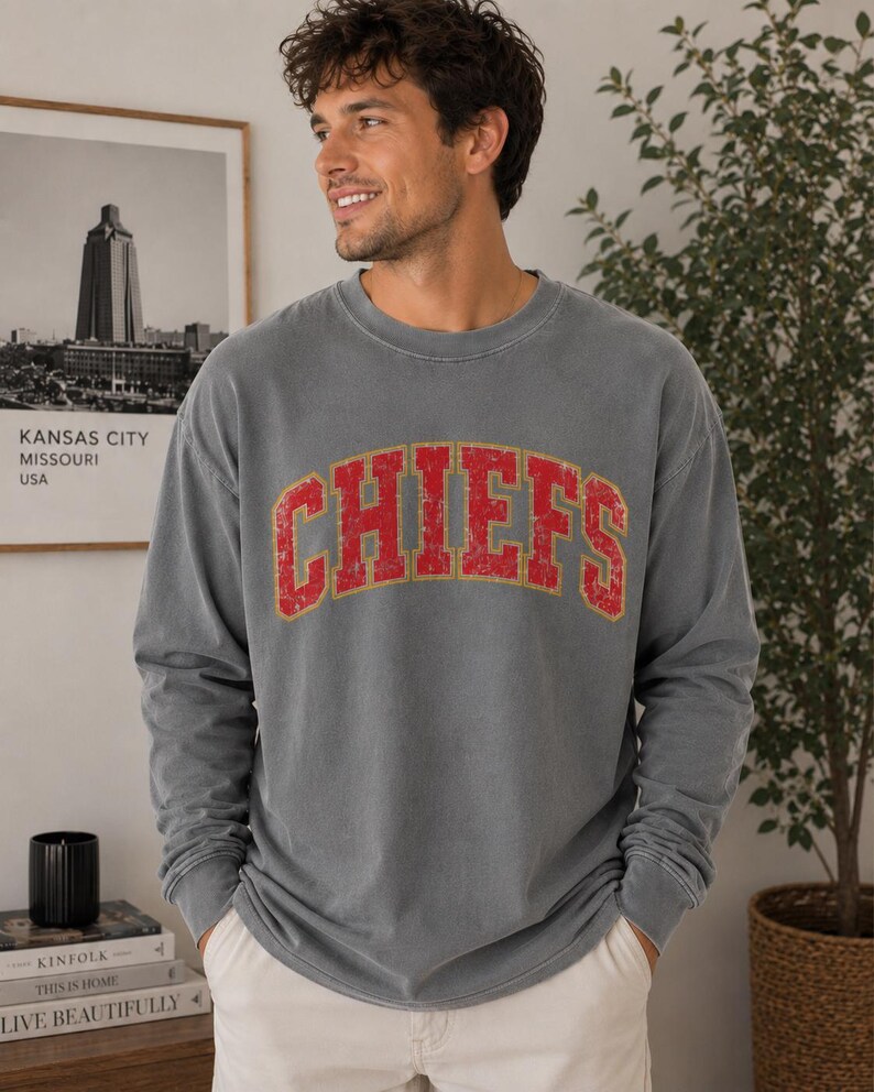 May include: A charcoal grey long-sleeve crewneck sweatshirt featuring the word CHIEFS printed in a distressed red font with a gold outline across the chest. The garment has a relaxed fit with ribbed cuffs and a ribbed neckline.