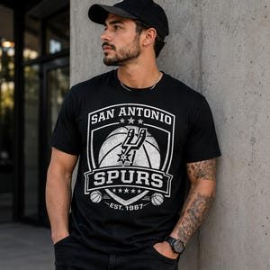 May include: Black t-shirt featuring the San Antonio Spurs logo in white. The design includes a basketball, the team name, and the establishment year, 1967. The shirt is short-sleeved and has a crew neck.