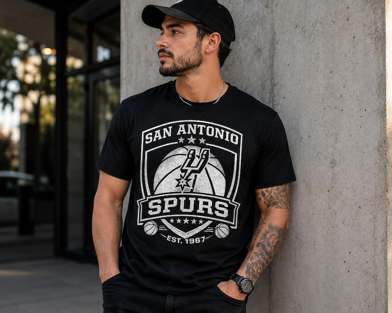 May include: Black t-shirt featuring the San Antonio Spurs logo in white. The design includes a basketball, the team name, and the establishment year, 1967. The shirt is short-sleeved and has a crew neck.