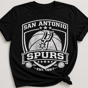May include: Black t-shirt featuring the San Antonio Spurs logo in white. The logo includes a basketball, the team name, and the year 1967. The shirt has a crew neck and short sleeves.