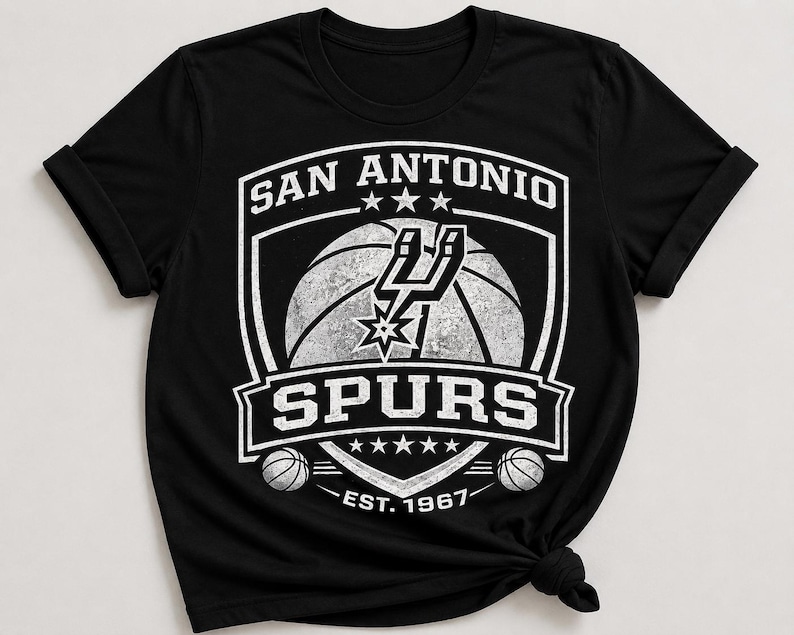 May include: Black t-shirt featuring the San Antonio Spurs logo in white. The logo includes a basketball, the team name, and the year 1967. The shirt has a crew neck and short sleeves.