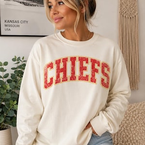 May include: A cream-colored long-sleeve crewneck sweatshirt featuring the word CHIEFS printed in a distressed red font with a yellow outline across the chest. The garment has a relaxed fit with ribbed cuffs and a ribbed neckline.