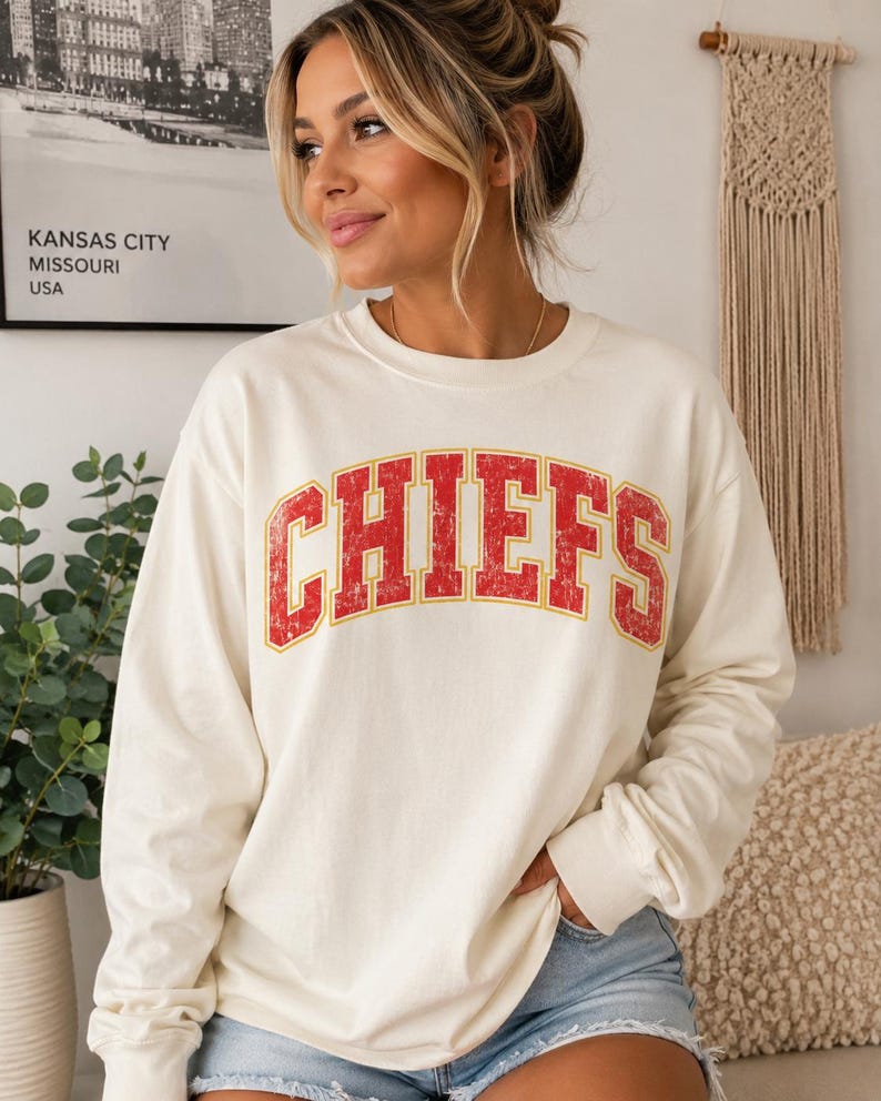 May include: A cream-colored long-sleeve crewneck sweatshirt featuring the word CHIEFS printed in a distressed red font with a yellow outline across the chest. The garment has a relaxed fit with ribbed cuffs and a ribbed neckline.