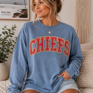 May include: A blue long-sleeve crewneck sweatshirt featuring the word CHIEFS printed in a distressed red font with a yellow outline across the chest. The garment has a relaxed fit with ribbed cuffs and a ribbed neckline, made of a soft fabric.