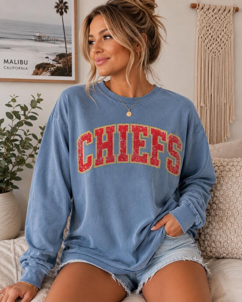 May include: A blue long-sleeve crewneck sweatshirt featuring the word CHIEFS printed in a distressed red font with a yellow outline across the chest. The garment has a relaxed fit with ribbed cuffs and a ribbed neckline, made of a soft fabric.