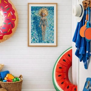 May include: A framed print of a doll in a pool, with a blonde hairstyle and sunglasses, wearing a black and white swimsuit. The image is surrounded by a red donut-shaped inflatable and a watermelon-shaped inflatable. A basket of toys and beach accessories are in the foreground.