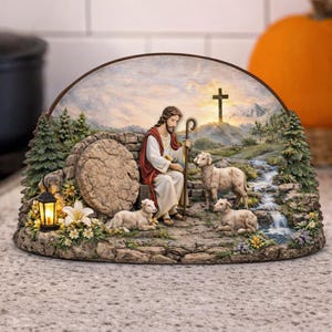 May include: A religious figurine depicting a biblical scene. It features a figure in white and red robes seated with a staff, surrounded by lambs. A cross is visible in the background, with a waterfall and trees. A lantern and flowers are also present.