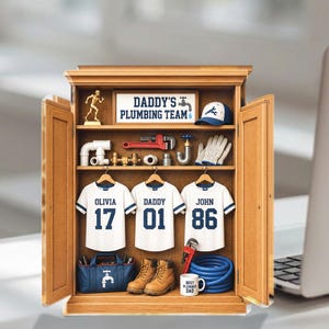 May include: A wooden cabinet with open doors displays plumbing tools, a baseball cap, work gloves, and three white t-shirts with blue accents. The shirts read "OLIVIA 17", "DADDY 01", and "JOHN 86". A sign reads "DADDY'S PLUMBING TEAM".