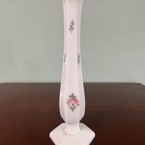 May include: A white, vintage-style vase with a hexagonal base and a ruffled top. The tall, slender vase features hand-painted pink and blue floral accents. The vase is made of opaque glass and stands on a dark wooden surface.