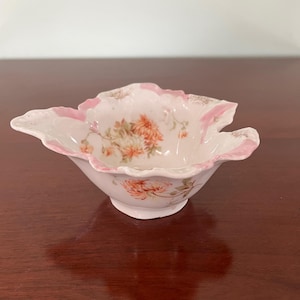 以下が含まれることがあります： 小さなアンティーク磁器の皿。縁はスカラップで、花の模様があります。皿は主に白で、ピンクのトリムがあり、オレンジとピーチ色の花が特徴です。この皿は、少量の料理を出すために使用される可能性があります。