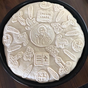May include: A round loaf of uncooked dough in a black baking pan, decorated with religious symbols. The decorations include crosses, wheat stalks, a dove, a lamb, grapes, and a central medallion with a saint. The text "СРЕЧНА СЛАВА" is also present.