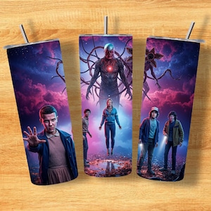 Pode incluir: Três canecas de aço inoxidável com canudos, cada uma com um design vibrante de uma série de ficção científica. A arte inclui personagens e criaturas em um cenário de céus roxos e azuis. As canecas são cilíndricas e têm tampas prateadas.