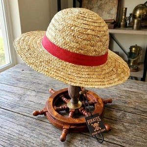 Puede incluir: Un sombrero de paja con una banda roja, exhibido en un soporte de rueda de barco de madera. El sombrero tiene un ala ancha y una textura tejida. Una etiqueta dice "Pirates Straw Hat Crew".