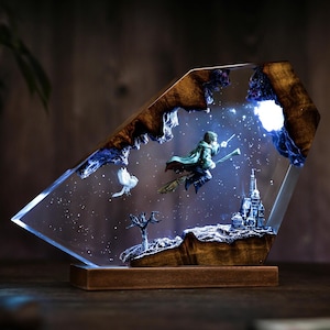 May include: A decorative resin sculpture featuring a wizard on a broomstick, a castle, and a bird, set against a starry night sky. The sculpture is encased in a clear, angled resin block with a wooden base and accents.