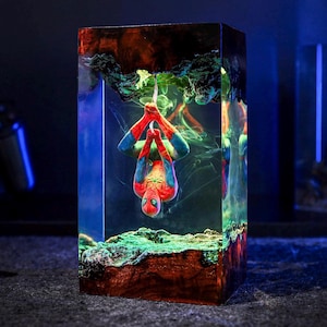 May include: A rectangular resin sculpture featuring Spider-Man hanging upside down. The sculpture has a vibrant color palette with red, blue, green, and orange hues. The base and top have a textured appearance, and the background has a smoky effect.