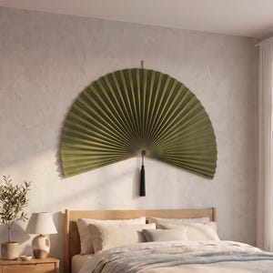 Puede incluir: Un abanico plegable grande y semicircular montado en la pared, de color verde oliva sólido. El abanico presenta una textura plisada y está suspendido de un pequeño gancho en la parte superior. Una borla negra larga y fina cuelga verticalmente.