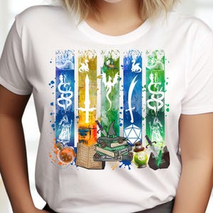 May include: White t-shirt with a fantasy-themed graphic. The design features vertical stripes with illustrations of swords, potions, books, and a wizard's hat. The artwork has a watercolor effect with paint splashes. The design is detailed and colourful.