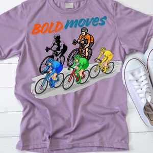 May include: A lilac t-shirt with a cycling graphic. The design shows cyclists in different coloured outfits, riding bicycles on a grey road. The words "BOLD moves" are printed across the top in orange and blue.