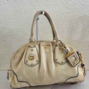 May include: Cream-colored leather handbag with gold-tone hardware. The bag features a top handle, a detachable tag, and the "PRADA" logo. The bag has a rounded shape and visible stitching details. The bag is sitting on a white surface.