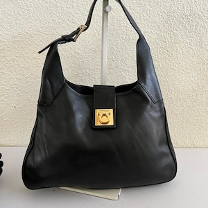 May include: Black leather shoulder bag with a single strap and gold-tone hardware. The bag features a rectangular flap closure with a gold-tone clasp and a large, curved body. The strap is adjustable with a gold buckle.