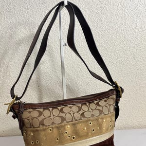 May include: A brown shoulder bag with a repeating logo pattern, gold accents, and a suede bottom. The bag has a long strap and a zipper closure. The bag is approximately 25 cm wide.