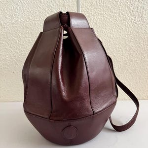 Op de afbeelding: Een donkerbordeauxleren bucket bag met een gestructureerd ontwerp uit meerdere panelen. De tas heeft een ronde onderkant, een ingesnoerde opening aan de bovenkant en een lange schouderriem. Een rond reliëflogo is zichtbaar op het voorpaneel.